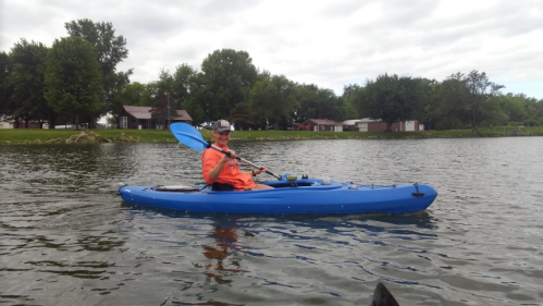 A person in an orange shirt paddles a blue kayak on a calm lake, surrounded by trees and a cloudy sky.