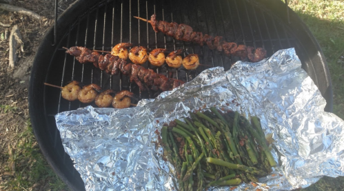 Grilled shrimp and beef skewers on a barbecue, with asparagus wrapped in foil beside them.
