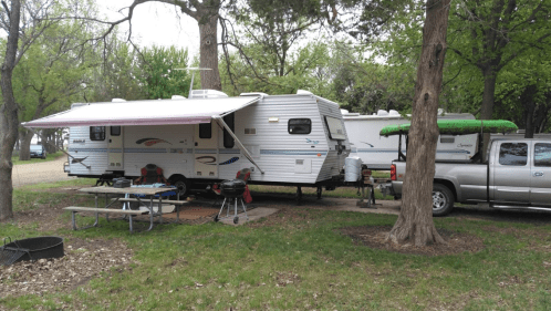 A camper with an awning parked beside a truck, surrounded by trees and a picnic area with a grill and table.