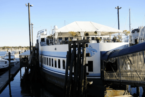 A large boat restaurant docked at a marina, featuring a white tent and decorative plants on the upper deck.