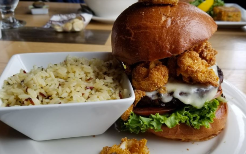 A gourmet burger topped with crispy shrimp, lettuce, and cheese, served with a side of seasoned rice.