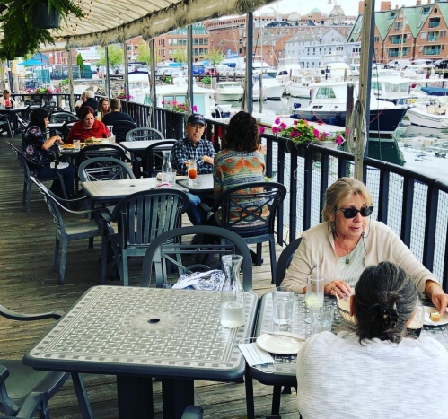 Outdoor dining area by a marina, with people at tables enjoying meals and drinks, surrounded by boats and flowers.