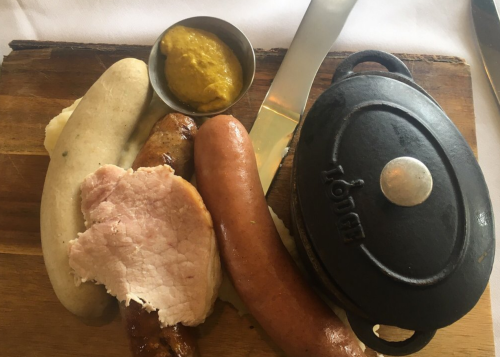 A wooden board with various sausages, sliced ham, mustard in a small bowl, and a cast iron pot.