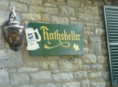 Sign for "Rathskeller" with a beer mug illustration, mounted on a stone wall beside a lantern and window shutters.