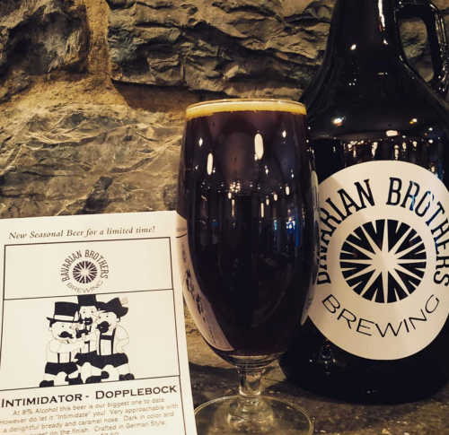 A dark beer in a glass next to a growler and a flyer for "Intimidator - Dopplebock" from Bavarian Brothers Brewing.