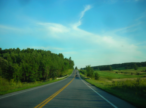 A scenic road stretches through green fields and trees under a blue sky with wispy clouds.