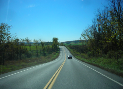 A winding road stretches into the distance, flanked by green fields and trees under a clear blue sky.