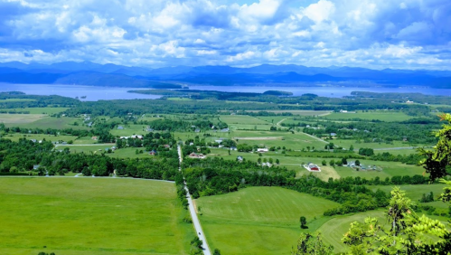 A panoramic view of lush green fields, a winding road, and a lake surrounded by mountains under a blue sky with clouds.
