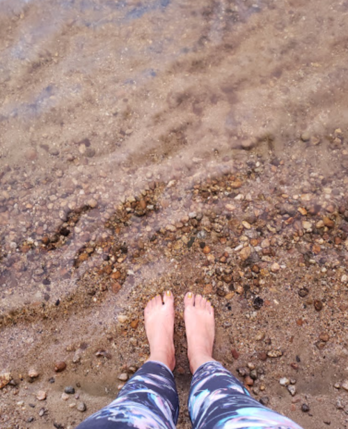 Person's feet standing on a rocky shore with clear water gently lapping at the edges.