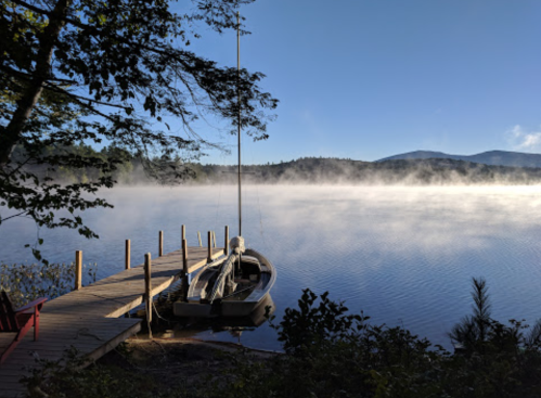 A serene lake scene with a dock, a boat, and mist rising from the water, surrounded by trees and mountains.