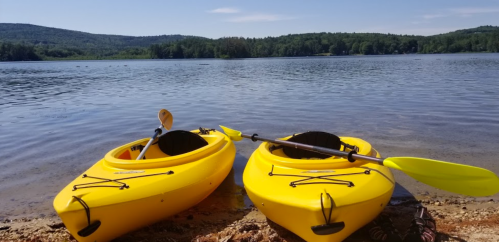 Two yellow kayaks on a sandy shore by a calm lake, surrounded by green hills under a clear blue sky.