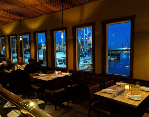 Cozy restaurant interior with tables set for dinner, featuring large windows overlooking a marina at dusk.