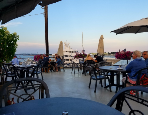 Outdoor dining area with tables, flowers, and a view of a marina at sunset. People are enjoying their meals.