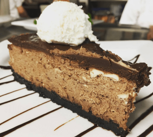 A slice of chocolate cheesecake with a dollop of whipped cream on top, served on a white plate with chocolate drizzle.