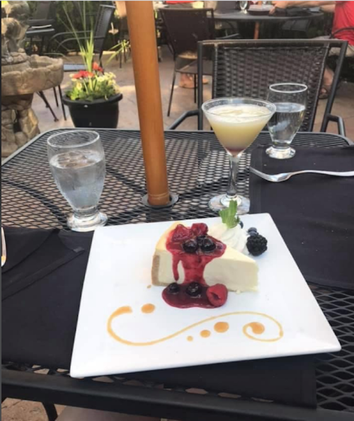 A slice of cheesecake topped with berries on a table with a cocktail and water glasses in a restaurant setting.