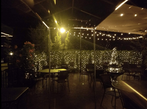A cozy outdoor dining area at night, illuminated by string lights and featuring a fountain surrounded by greenery.