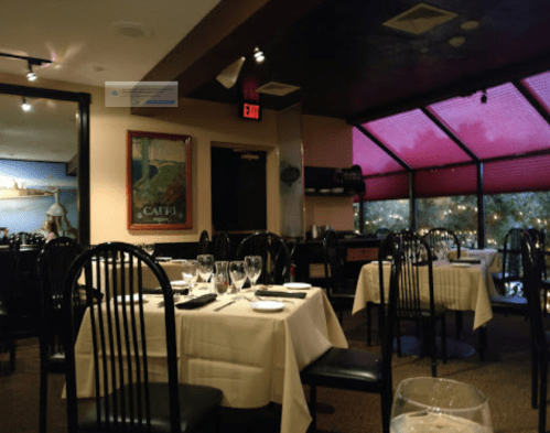 A cozy restaurant interior with tables set for dining, featuring black chairs and large windows with red shades.