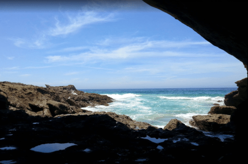 View of the ocean from a rocky cave, with waves crashing against the shore under a clear blue sky.