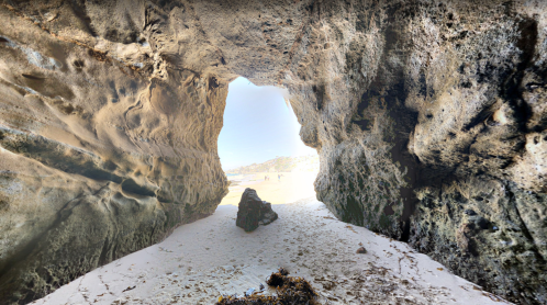 View from inside a rocky cave, looking out at a sandy beach and distant shoreline under a bright sky.