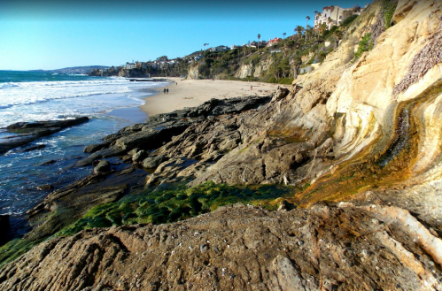 A scenic beach view with rocky shoreline, gentle waves, and people walking along the sand under a clear sky.