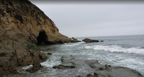 Rocky coastline with a cave, gentle waves lapping at the shore under a cloudy sky.