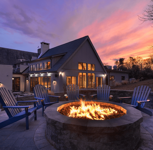 A cozy outdoor fire pit surrounded by blue chairs, with a modern house and colorful sunset in the background.