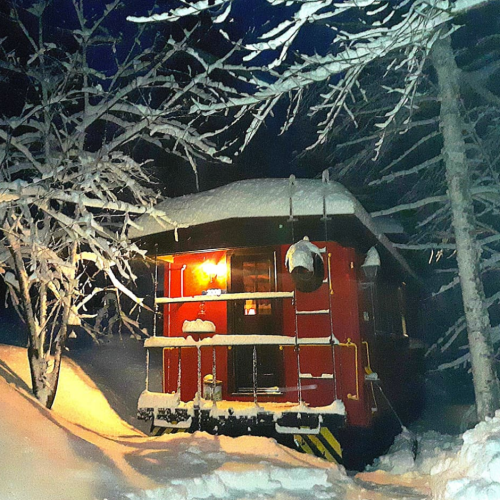 A cozy red train car illuminated at night, surrounded by snow-covered trees and a blanket of fresh snow.