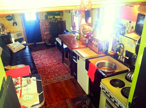 Cozy kitchen and living area with a black sofa, wooden table, and colorful rugs in a small, well-lit space.