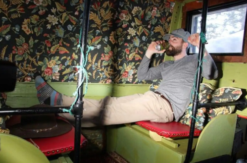 A man relaxes in a colorful, floral-patterned interior, sipping a drink with his feet up on a seat.