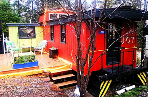 A colorful tiny house with a red exterior, surrounded by trees and outdoor seating, illuminated by string lights.
