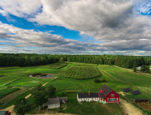Aerial view of a red farmhouse surrounded by green fields, trees, and a pond under a cloudy blue sky.