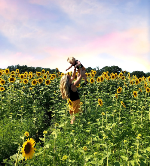 A woman holds a child in a sunflower field, surrounded by vibrant yellow blooms under a colorful sky.