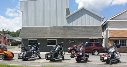 A row of motorcycles parked in front of a building with a sign that reads "Joey's." Clear blue sky in the background.