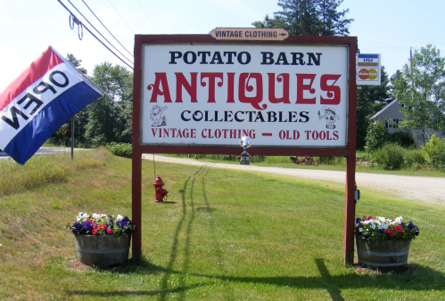 Sign for "Potato Barn Antiques" featuring vintage clothing and old tools, with flowers and an "OPEN" flag nearby.