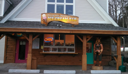 Exterior of Naughty Nellie's Ice Cream Bar, featuring a wooden sign, open window, and a carved statue outside.
