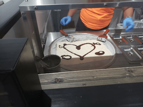A person in an orange shirt prepares rolled ice cream, creating a heart shape with chocolate sauce on a cold surface.