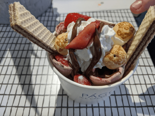 A cup of ice cream topped with whipped cream, strawberries, chocolate drizzle, popcorn, and wafer sticks.