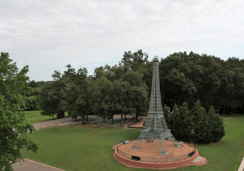 A miniature Eiffel Tower surrounded by trees in a grassy park setting under a cloudy sky.