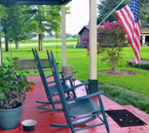 A porch with rocking chairs, a potted plant, and an American flag, surrounded by green grass and trees.