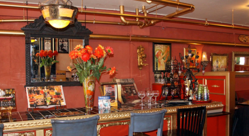 A stylish bar with a red wall, ornate decor, flowers, and various drinks displayed on the counter and shelves.