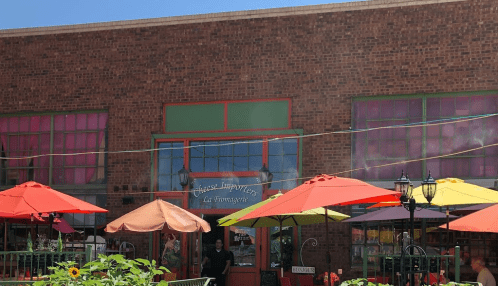 A brick building with colorful umbrellas outside, featuring a sign that reads "Cheese Importers." Bright and inviting atmosphere.