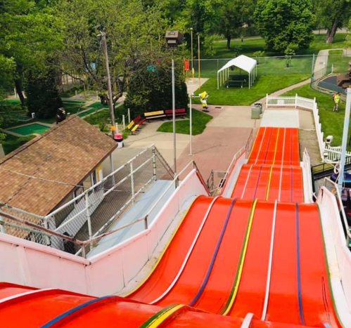 A colorful water slide with red and yellow lanes, surrounded by greenery and park amenities in the background.