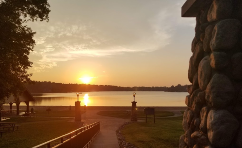 A serene sunrise over a calm lake, with a stone pathway and picnic area in the foreground.