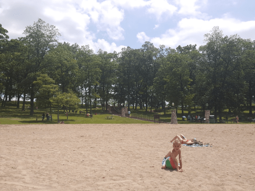 A sandy beach scene with a child playing with a beach ball, surrounded by trees and people in the background.