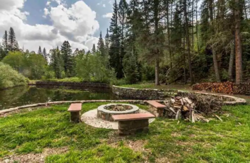 A serene outdoor setting with a stone fire pit, benches, and a pond surrounded by trees and stacked firewood.