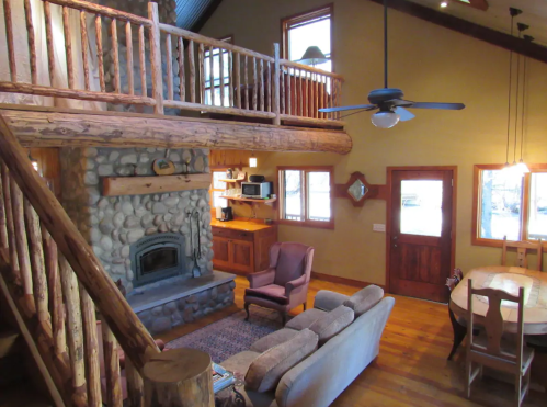 Cozy cabin interior with a stone fireplace, wooden beams, and a loft area, featuring a sofa and dining table.