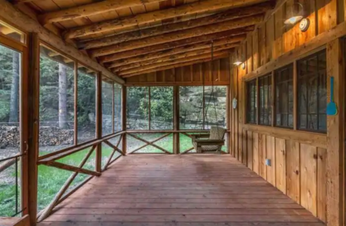 A wooden screened porch with a rustic design, featuring a bench and views of greenery outside.