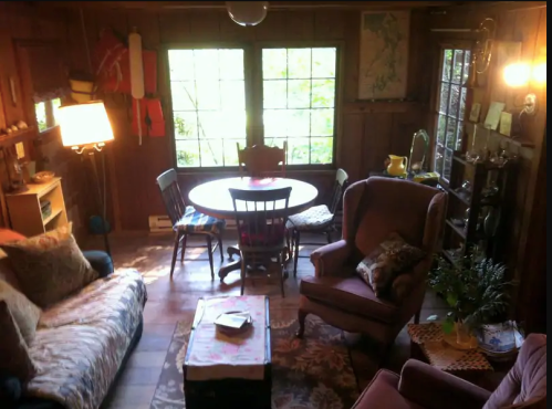 Cozy wooden living room with a round dining table, chairs, a sofa, and large windows letting in natural light.
