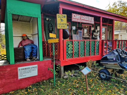 A colorful cabin with a sign for "Camp Goofbeer," featuring a mannequin sitting on the porch and a nearby sign.