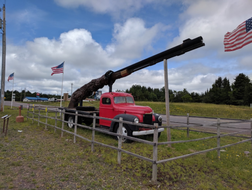 A red vintage truck with a large wooden cannon mounted on its bed, surrounded by American flags and a grassy area.